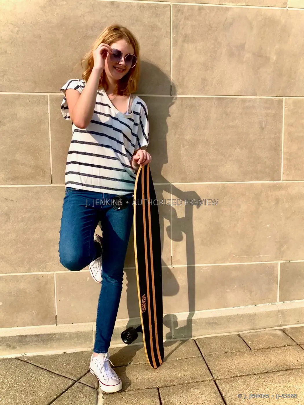 Young woman posing with a longboard against a sunlit stone wall